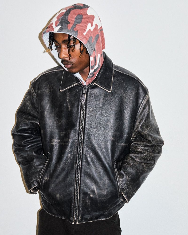Adult male wearing a black leather jacket and camouflage hoodie stands indoors against a white wall in a medium portrait shot.