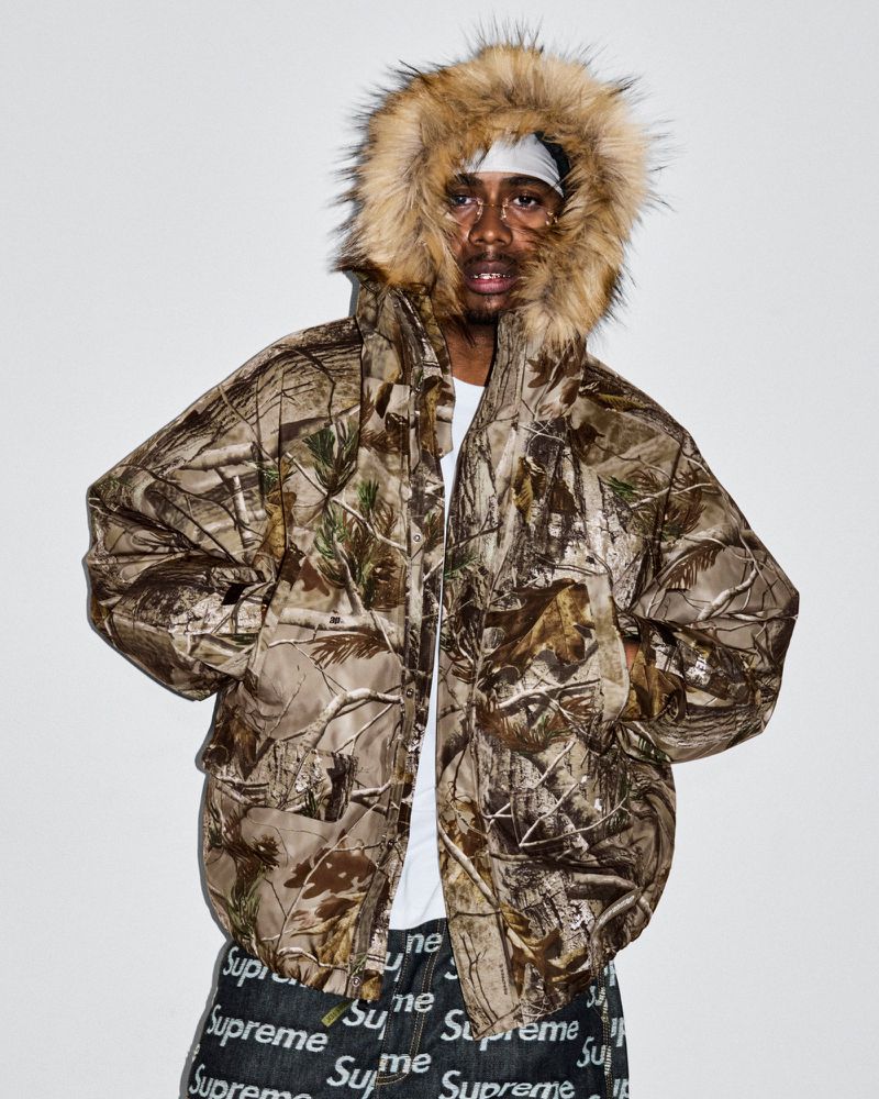 A man wearing a camouflage puffer jacket with a fur-lined hood is centered against a bright white backdrop. Fashionable street style.