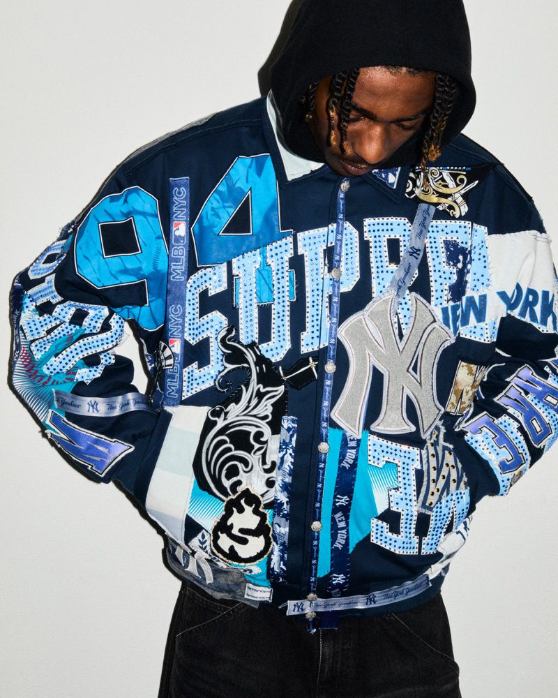 A person wearing a Supreme New York Yankees jacket. The jacket features light blue, white, and navy patterns and various appliqués.