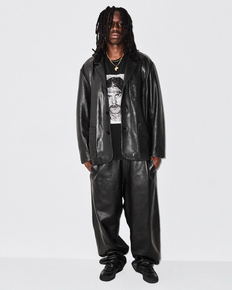 Man in a black leather suit with a graphic tee stands against a white backdrop in a studio shot with fashion style.