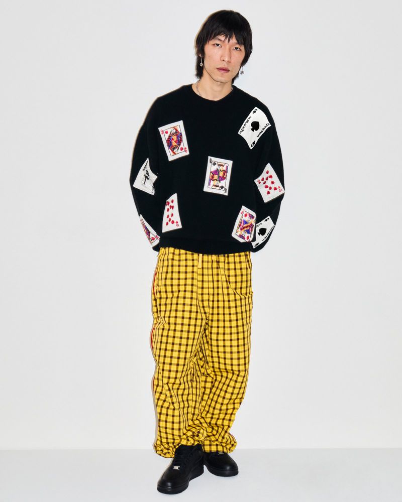Full-length portrait of a slender Asian adult male wearing a black sweater with playing card graphics and yellow plaid pants in a studio.
