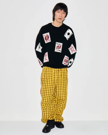 Full-length portrait of a slender Asian adult male wearing a black sweater with playing card graphics and yellow plaid pants in a studio.