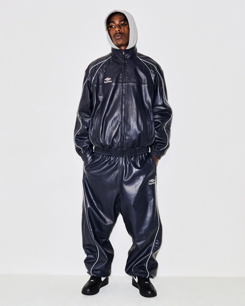 An African-American man in a blue faux leather tracksuit and black sneakers stands against a white background in a studio portrait.