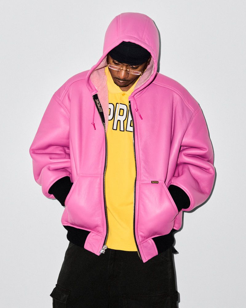 Man in pink leather hooded jacket, yellow t-shirt, and black pants poses against a white background in a studio shot.