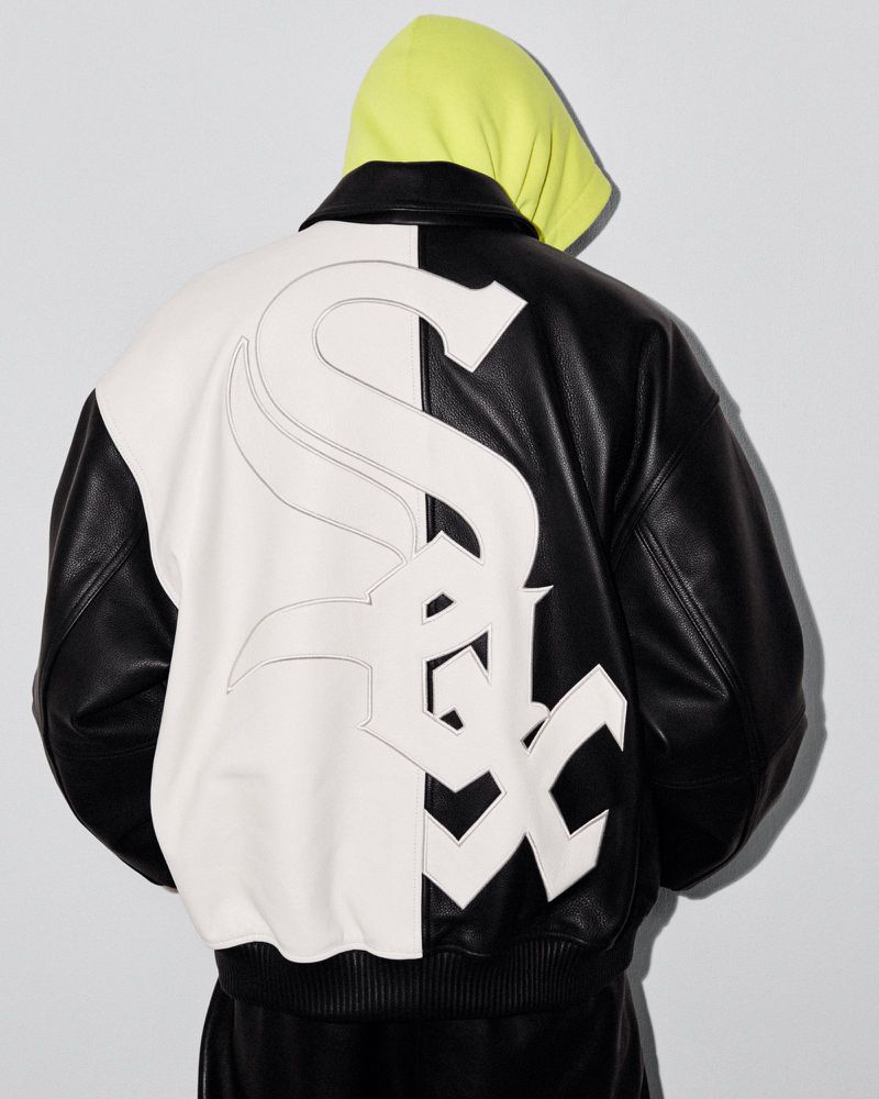 Person wearing a black and white Chicago White Sox leather bomber jacket with a yellow hoodie on a neutral background in a studio setting.
