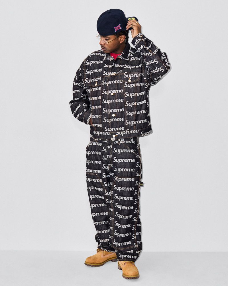 Adult male poses in a denim jacket and pants with a Supreme logo pattern, wearing a hat and glasses against a neutral background.