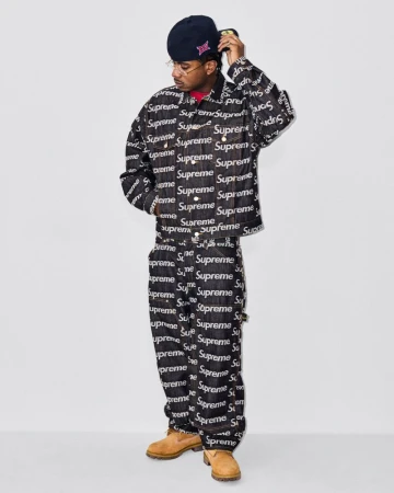 Adult male poses in a denim jacket and pants with a Supreme logo pattern, wearing a hat and glasses against a neutral background.
