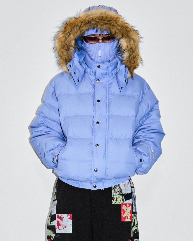 Person wearing a fur-trimmed, light-blue puffer jacket and patterned pants stands against a white backdrop in a studio setting.