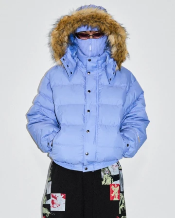 Person wearing a fur-trimmed, light-blue puffer jacket and patterned pants stands against a white backdrop in a studio setting.