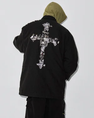 Rear view of a model wearing an olive hoodie and a black work jacket with an ornate cross graphic on the back against a white background.
