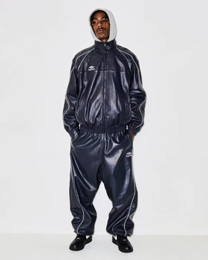 Man wearing a navy leather track jacket and matching track pants with white piping over a grey hoodie against a white background