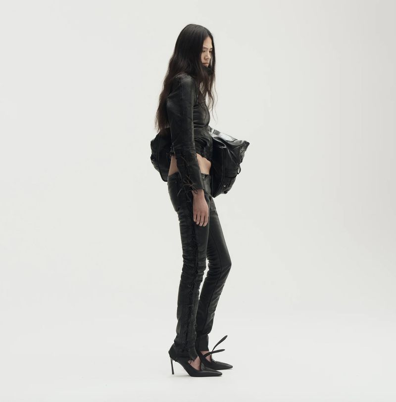 A slender woman with long dark hair poses in a black leather jacket, matching pants, and black heels against a bright white backdrop.