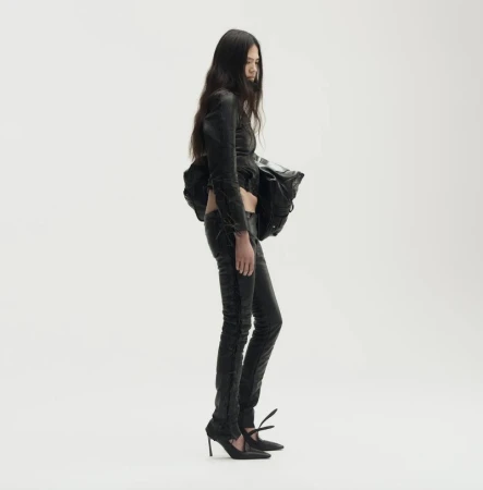 A slender woman with long dark hair poses in a black leather jacket, matching pants, and black heels against a bright white backdrop.