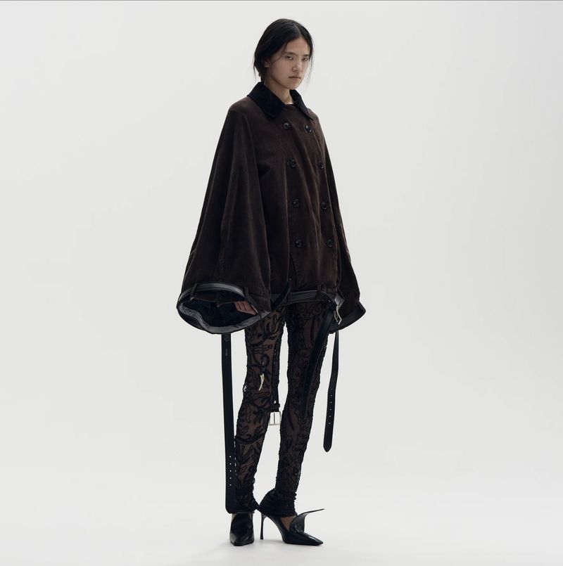 A fashion portrait features a slender young woman posing in a dark brown, oversized jacket and patterned tights against a bright white backdrop.