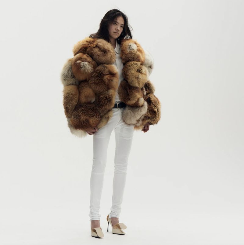 An Asian woman wearing a fur coat, white pants, and heeled shoes is standing in a bright white photo studio, fashion forward pose.