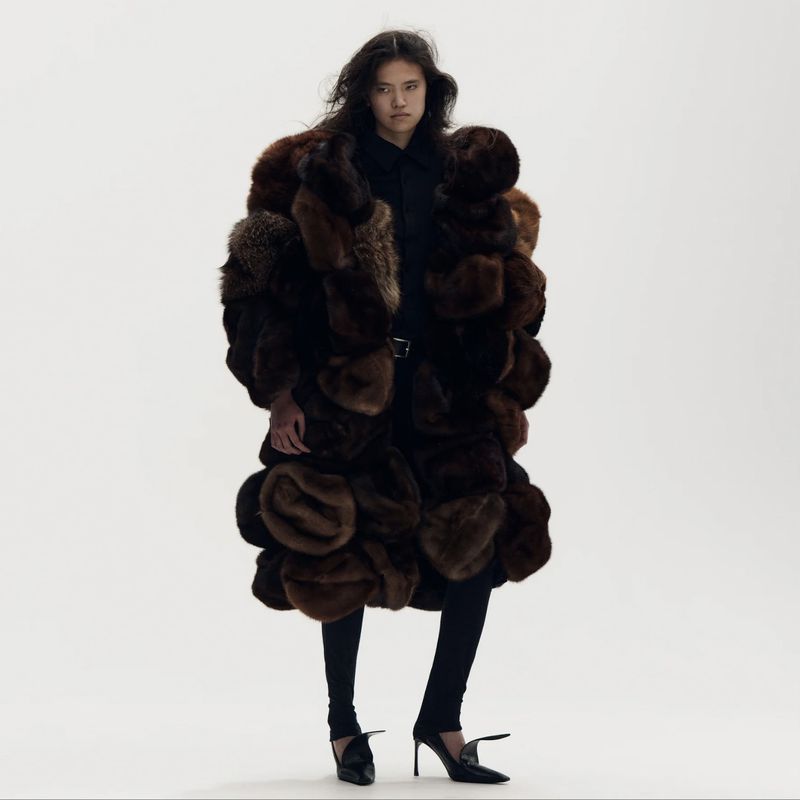 Model wearing an oversized sculptural brown patchwork fur coat with black trousers and heels against a white backdrop.