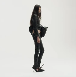 Model in all-black outfit with leather trousers and heels holding a large bag against a white background.