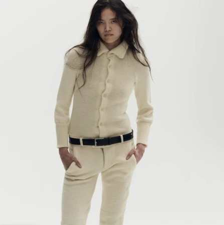 Model wearing an off-white knit collared top and matching knit trousers with a black belt against a white studio background.