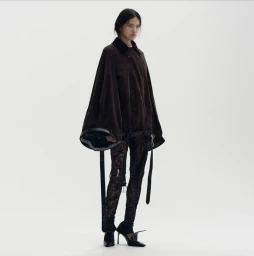 Model wearing an oversized dark brown cape jacket with hanging straps, patterned lace tights, and black pointed heels on a white backdrop.