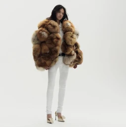 Model wearing a voluminous brown patchwork fur jacket, white slim trousers, and pointed heels against a white seamless backdrop.
