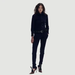 Model standing against a white seamless background wearing a dark buttoned top, trousers, belt, and heels for Hodakova.