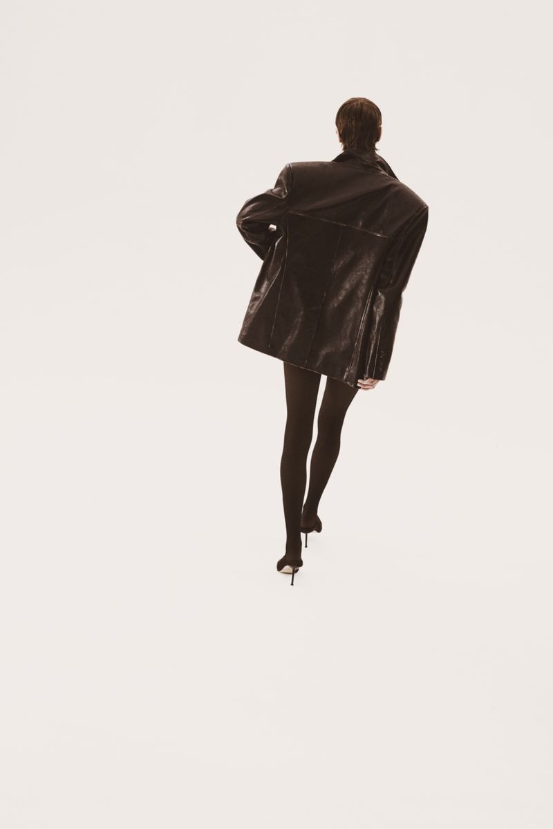 Adult female walking away in a black leather jacket, black tights, and high heels on a bright white studio backdrop; fashion editorial.