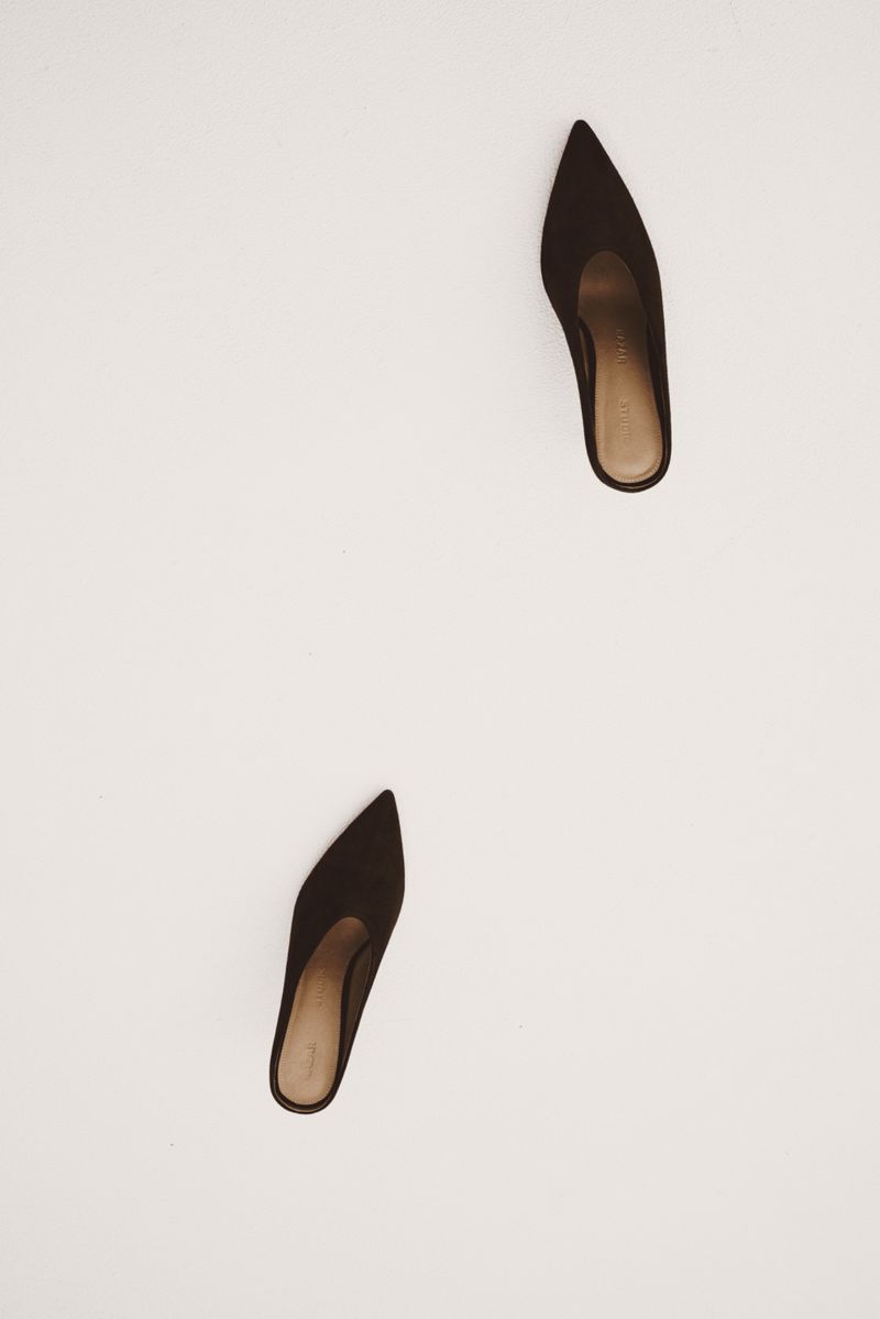 Pair of brown, pointed-toe heeled mules on a white surface, showcasing minimalist style and elegant footwear fashion.