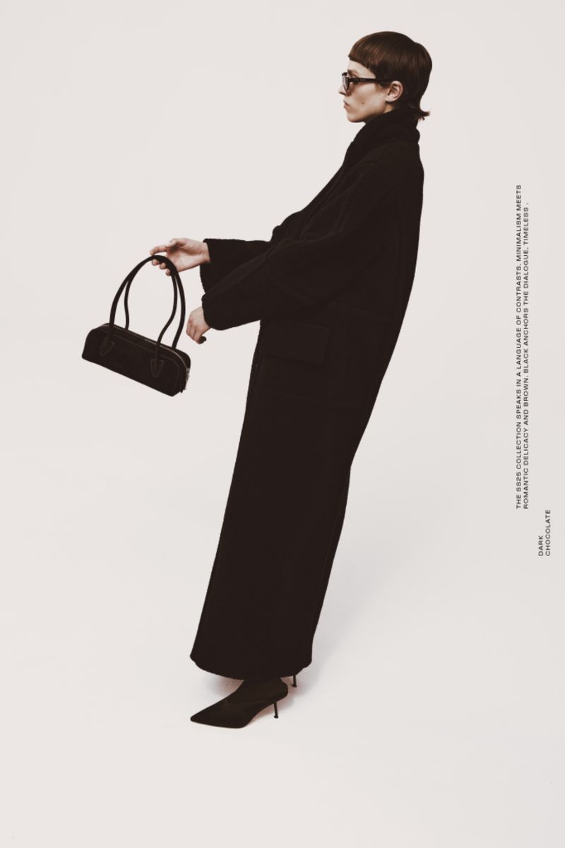 A woman in a long black coat poses in a studio, holding a dark handbag. The image has a fashion editorial style and studio lighting.
