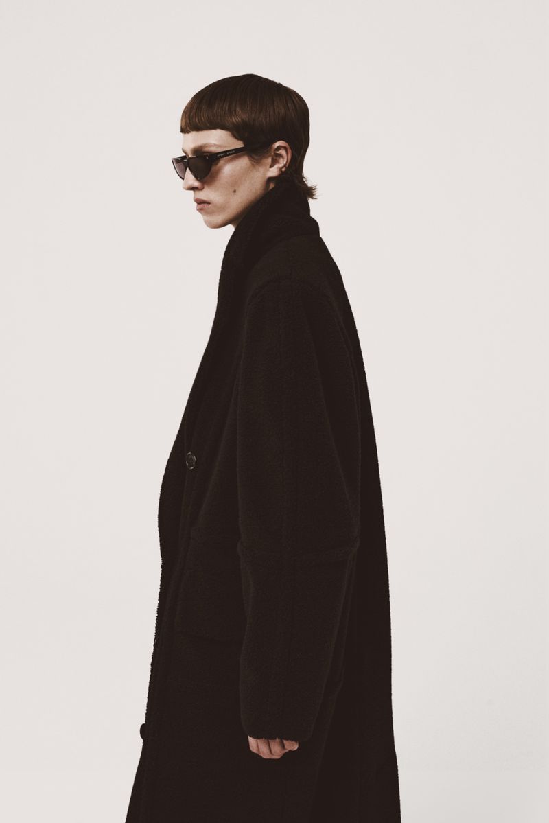 A person with a short haircut and sunglasses wearing a long black coat poses in a photo studio with a white background.