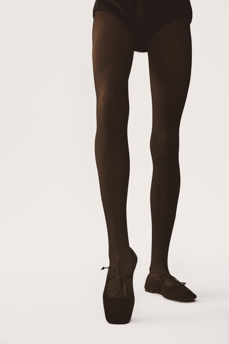 A close-up studio shot of a ballet dancer's legs and feet wearing black tights and ballet shoes against a white backdrop. Focus on pose.