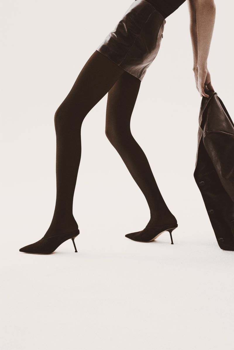 Fashion shot featuring a model wearing a mini skirt, black stockings, and high heel shoes, holding a jacket in a bright studio.
