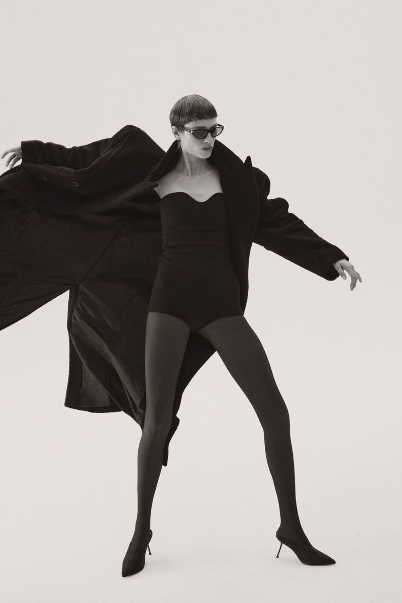 Fashion shot of a woman in a black bodysuit, tights, and heels, wearing sunglasses and a coat against a white background.