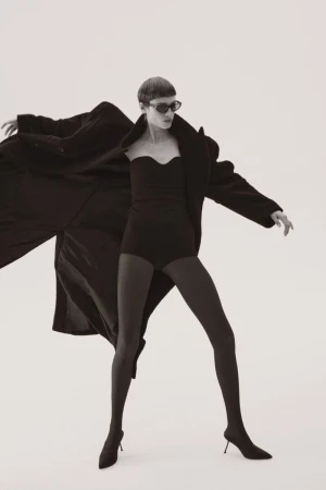 Fashion shot of a woman in a black bodysuit, tights, and heels, wearing sunglasses and a coat against a white background.