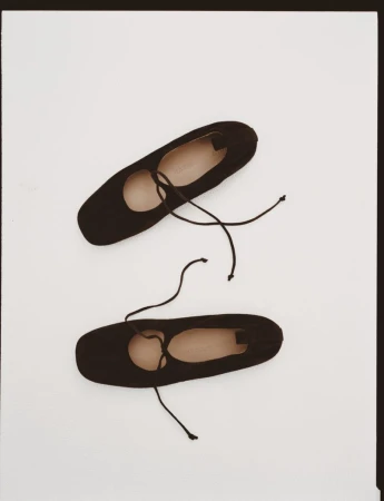 Two black ballet shoes, or pointe shoes, sit against a white background. The shoes are angled with laces and a plain design.