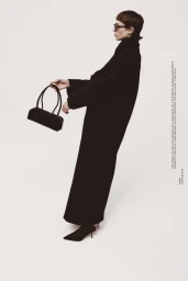 Model wearing a long black coat and holding a black handbag against a white background for Kazar Studio capsule collection.