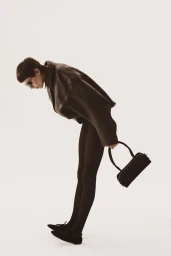 Model leaning forward against a white background wearing a brown leather jacket and holding a black handbag.