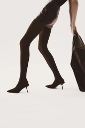 Model wearing dark tights and stiletto heels holding a leather jacket against a white background for Kazar Studio