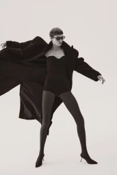Model wearing a strapless black bodysuit, opaque black tights, pointed heels, and an oversized black coat on a white studio backdrop.