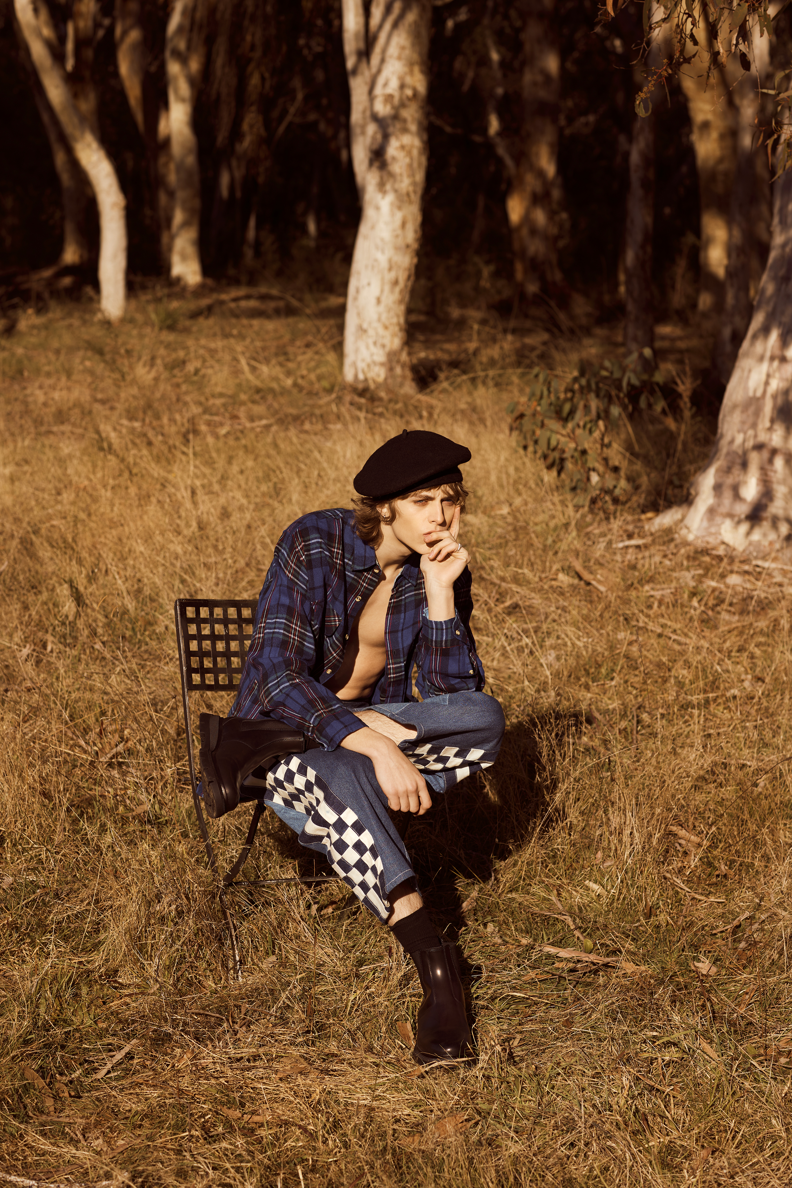 Young adult male in stylish outfit sits pensively on a chair in a sun-drenched field near trees, wearing plaid shirt and checkerboard pants.