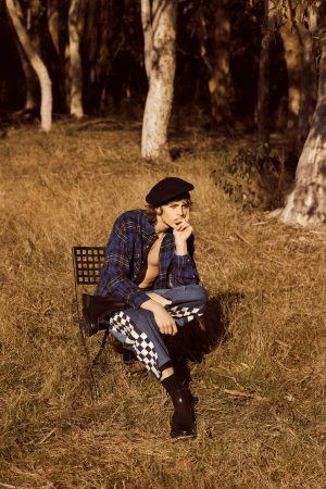 Young adult male in stylish outfit sits pensively on a chair in a sun-drenched field near trees, wearing plaid shirt and checkerboard pants.