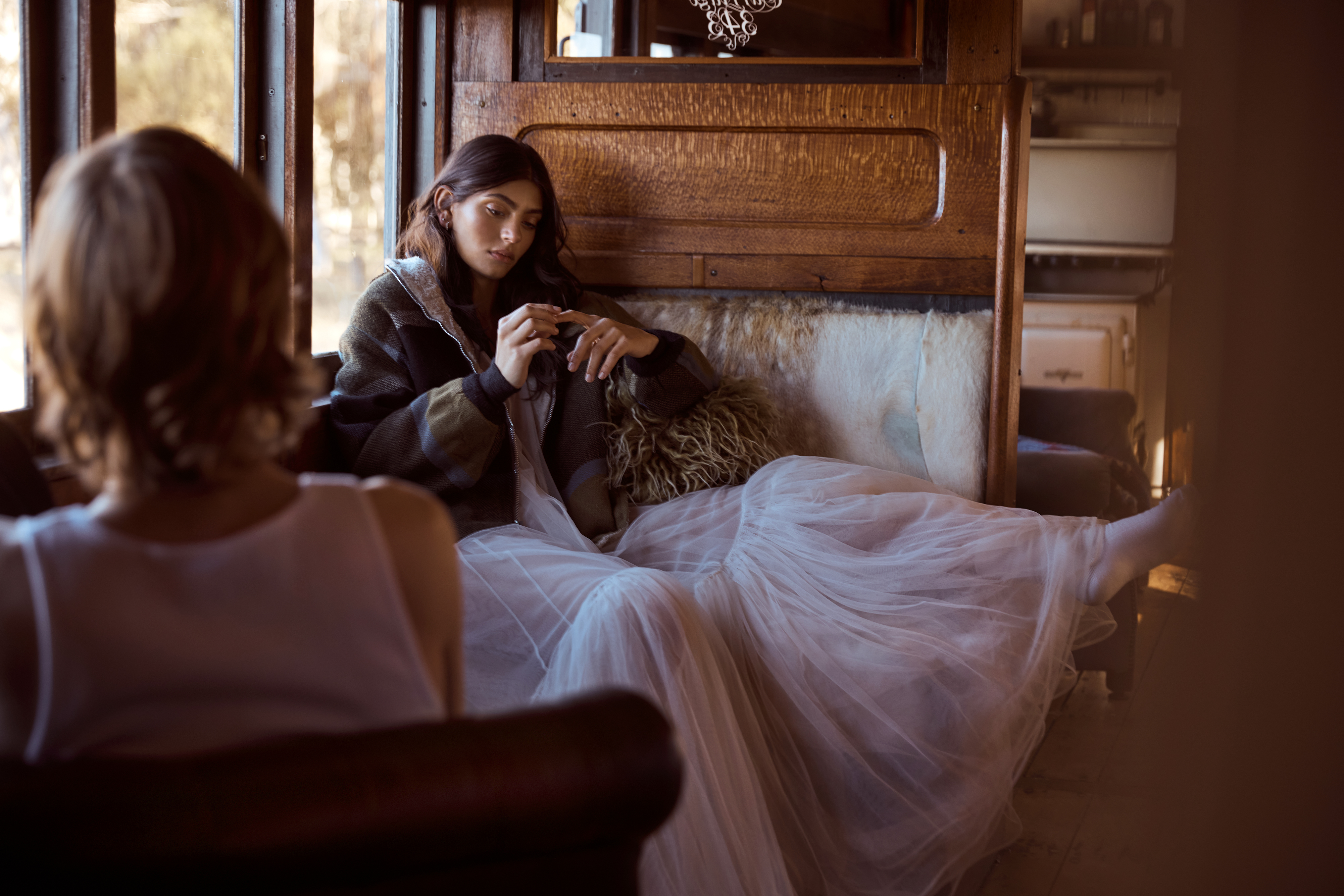 Indoor scene features a woman seated, wearing a tulle skirt and jacket, while a second woman is seated in the foreground.