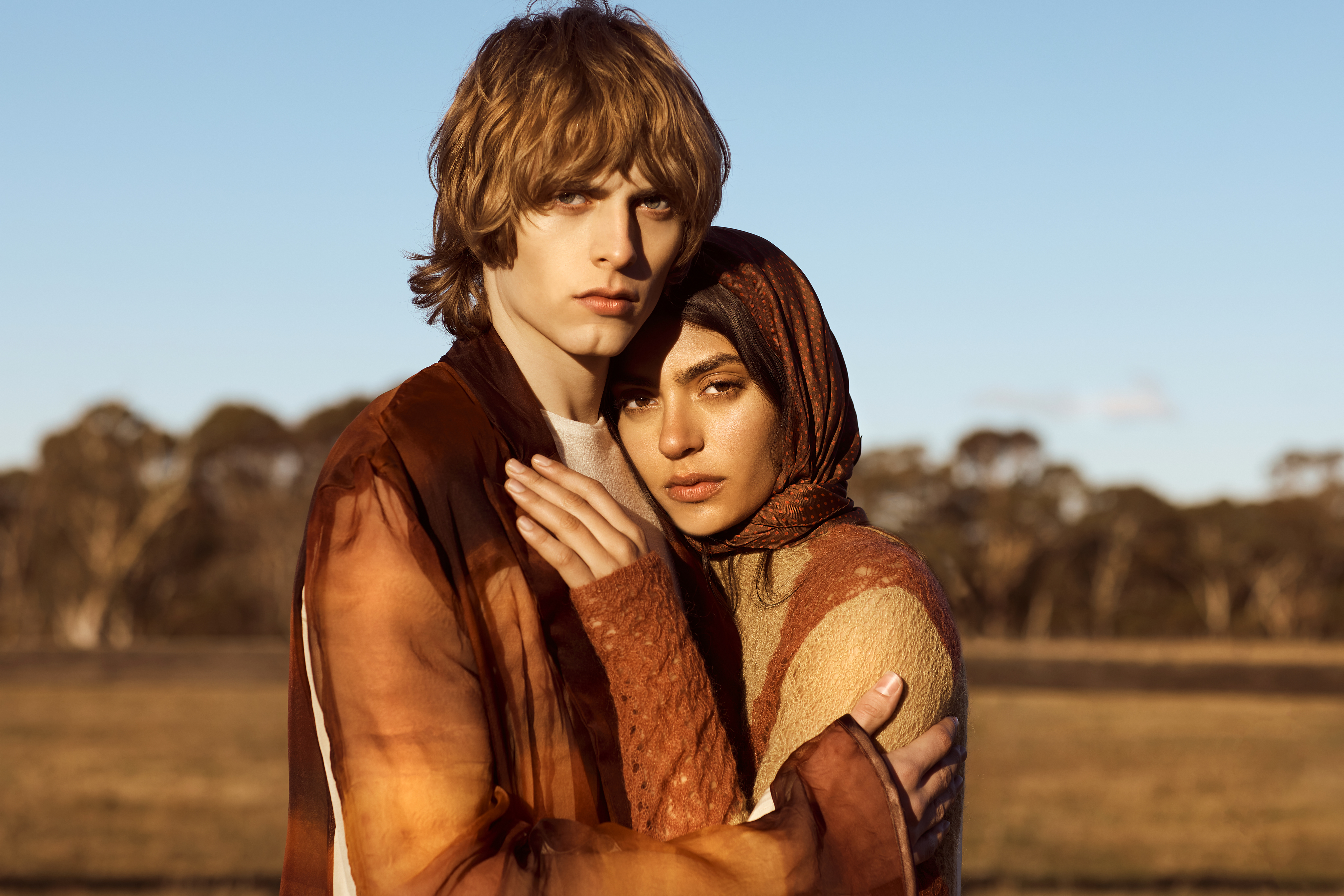 A young adult couple in an outdoor fashion shot. The man and woman are closely posed, with a field and trees in the background.