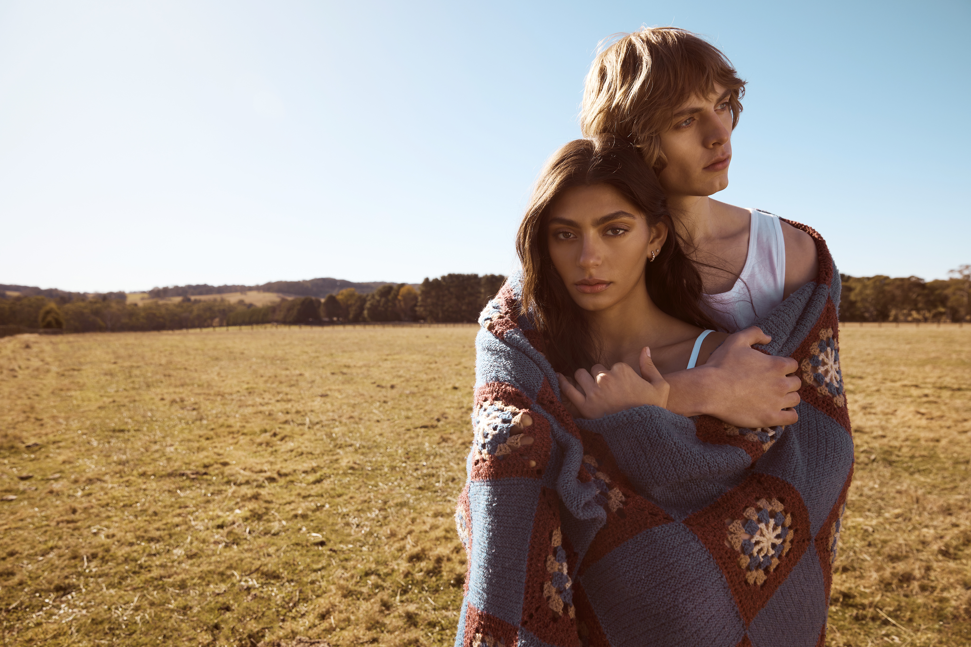 Young couple wrapped in a crocheted blanket standing in a field on a sunny day with a background of rolling hills and blue sky.