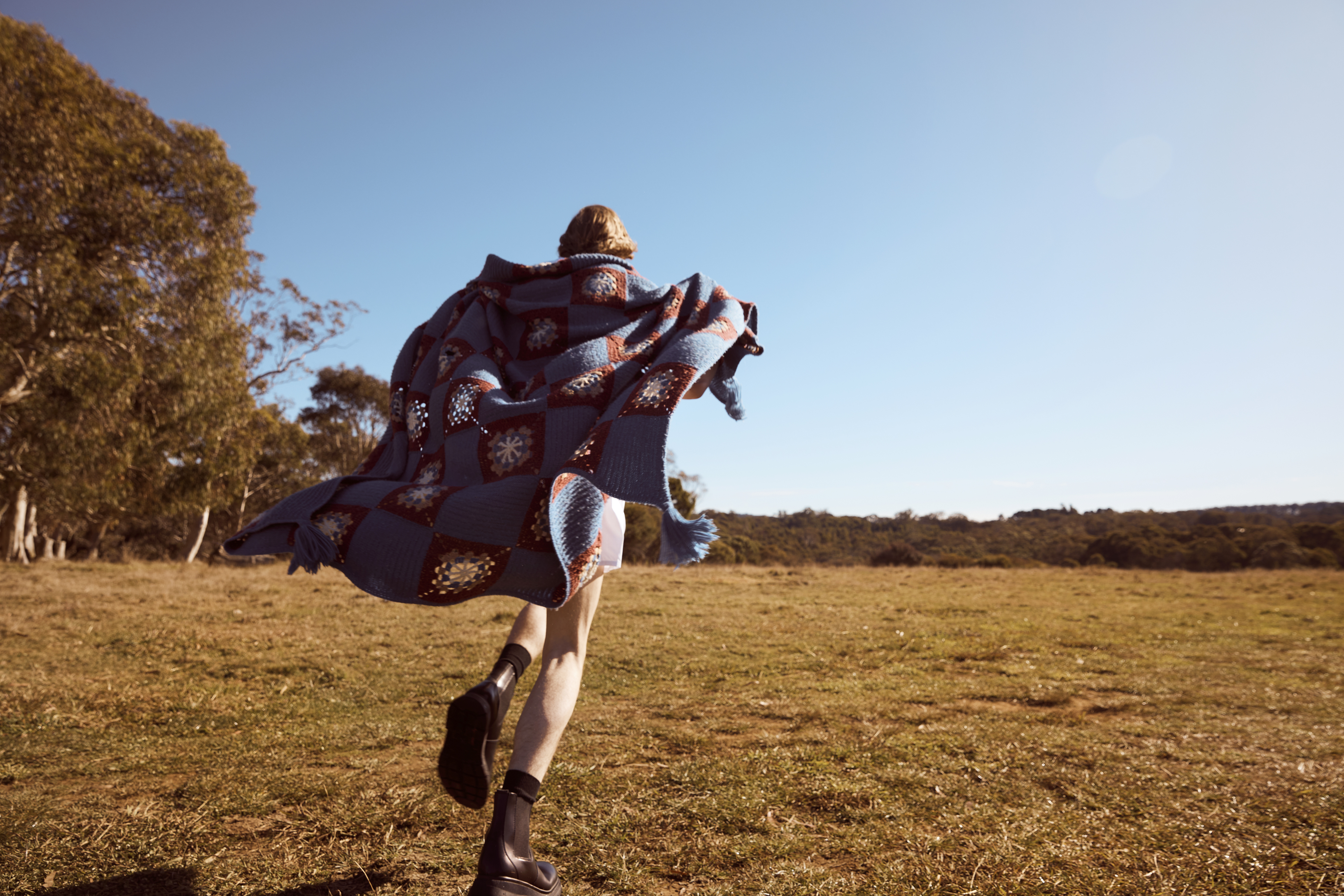 A woman with a crocheted blanket runs in a field of dry grass under a clear blue sky, wearing black boots and short white shorts.