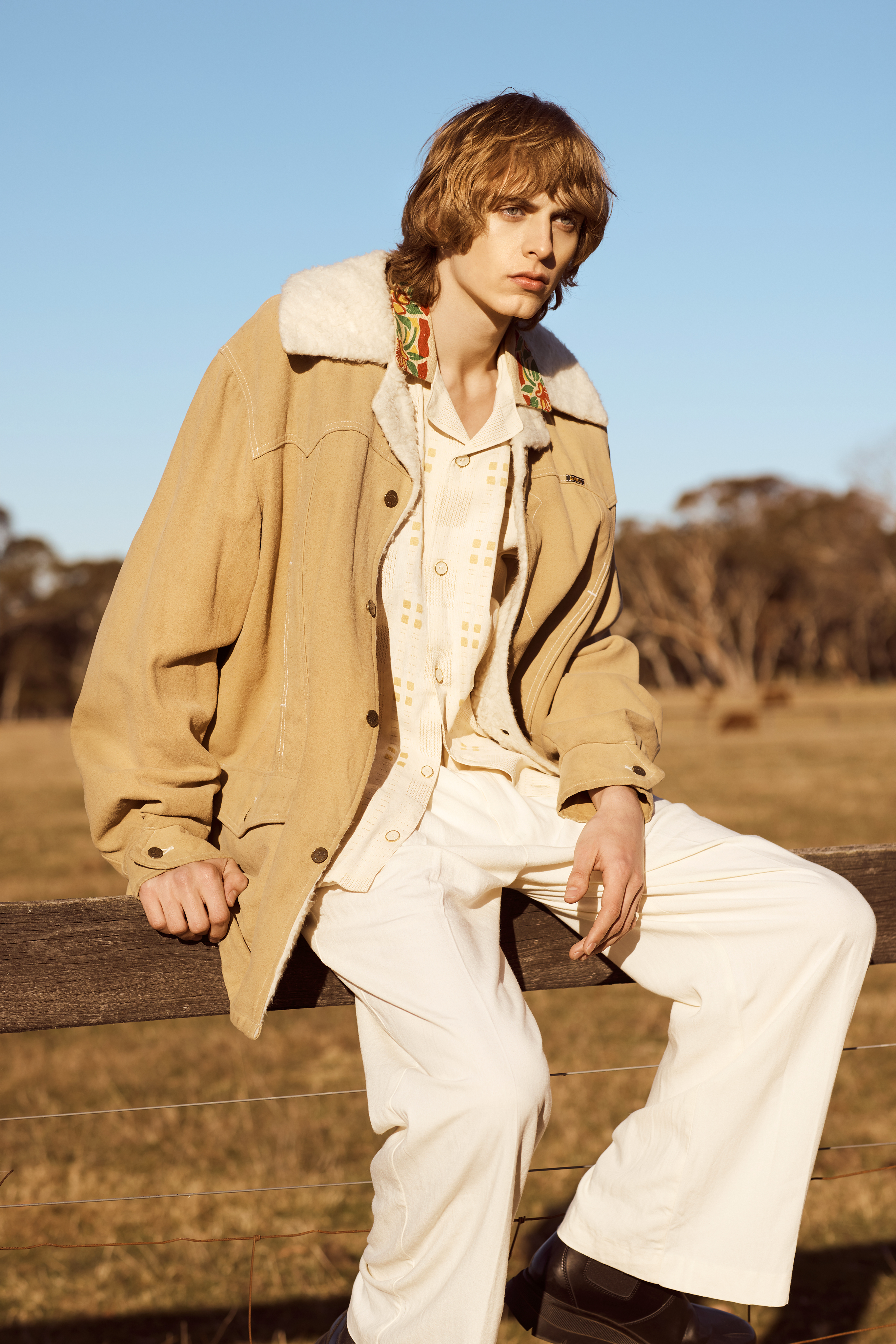 An adult male model poses in an outdoor fashion shot wearing a tan jacket, cream-colored pants, and platform shoes sitting on a fence.