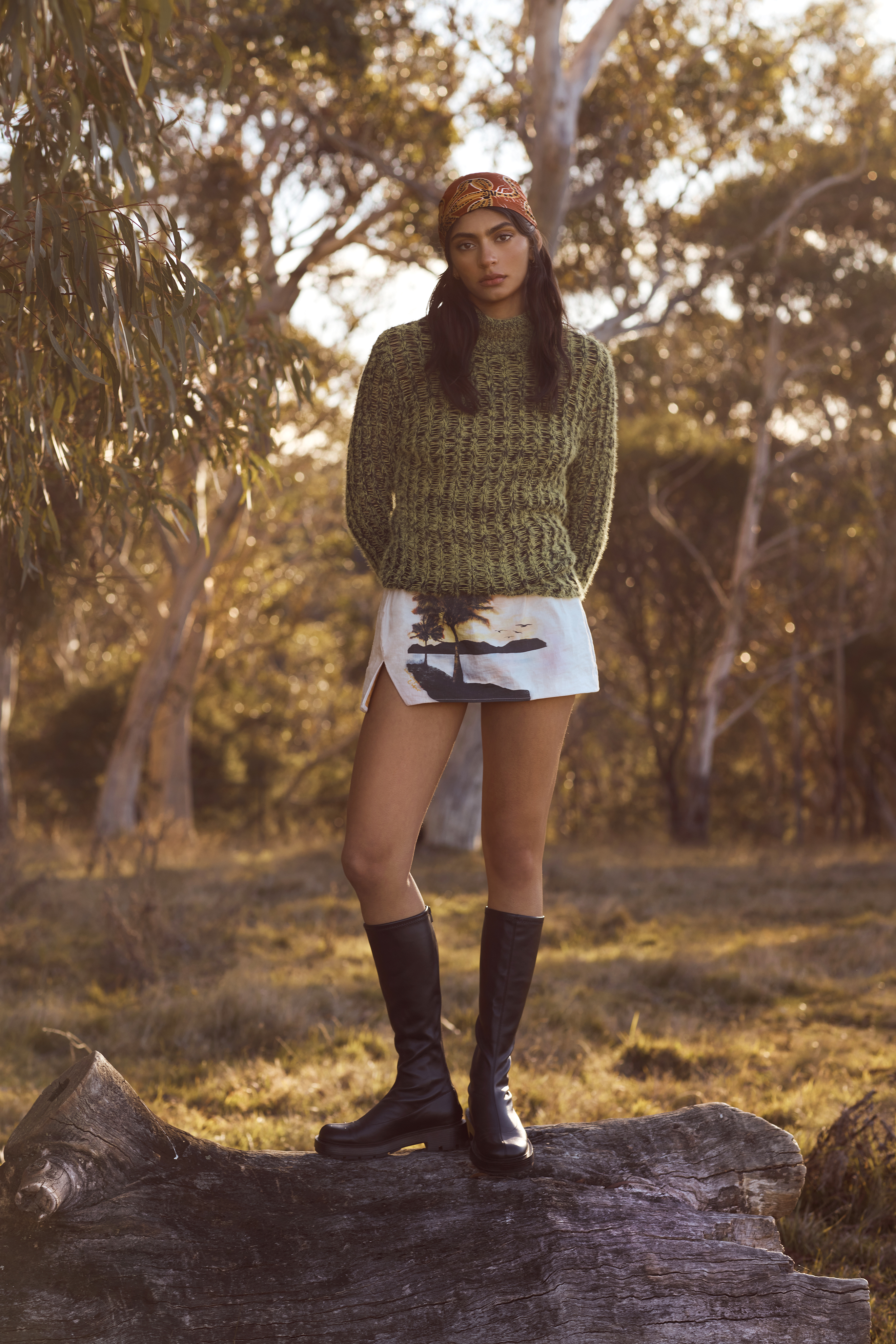 A woman in a knit sweater, shorts, and boots stands on a log in a field with trees in the background during golden hour.