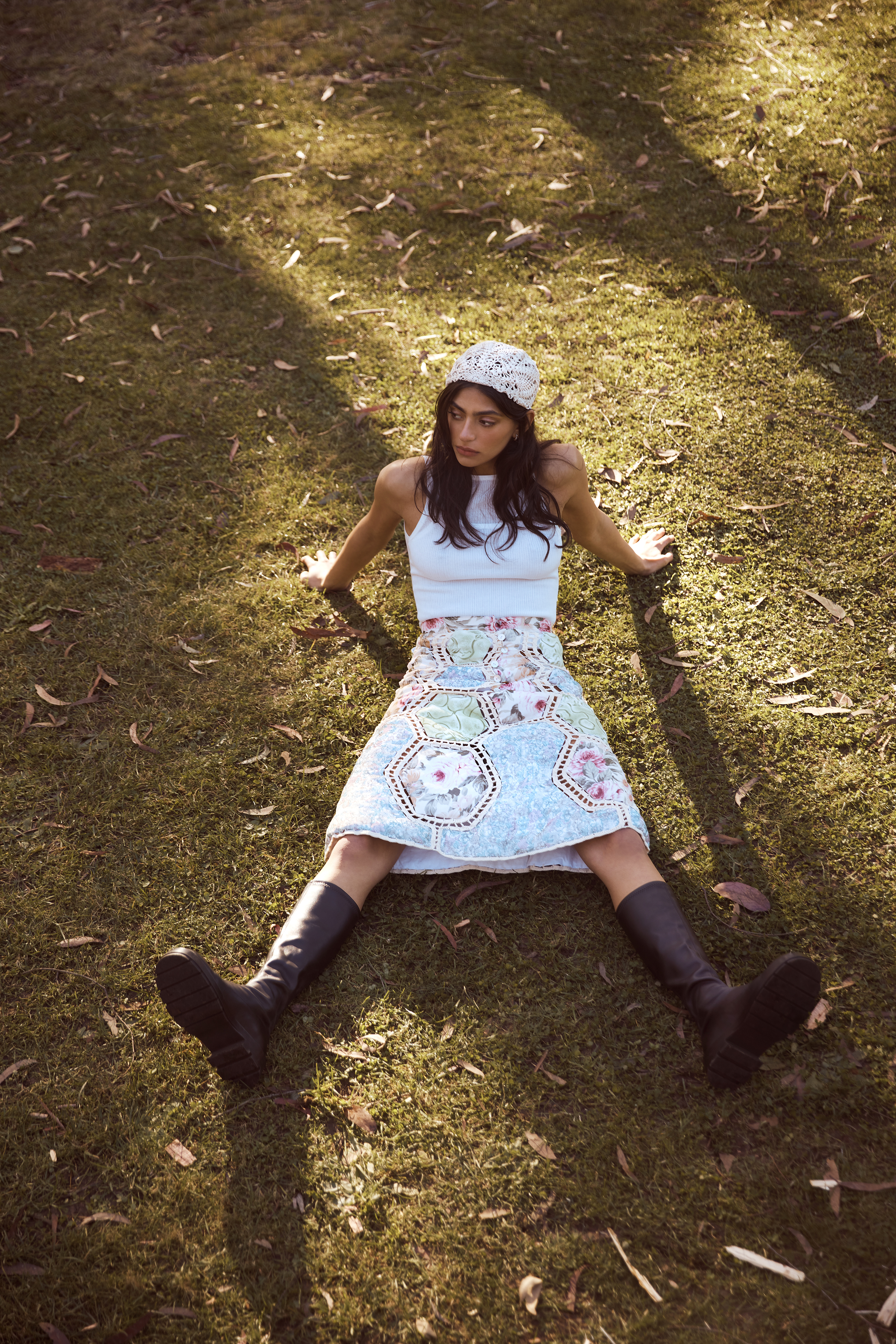 Woman in white top and floral skirt sits on grass in sunlight wearing black boots and white head covering, angled portrait.