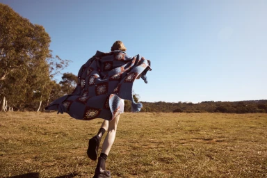 Model in blue crocheted blanket cape running across grassy field under clear sky