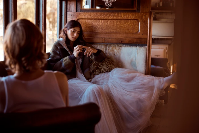 Two women inside a wooden train car, one seated on a plush couch wearing a tulle skirt and patterned jacket.