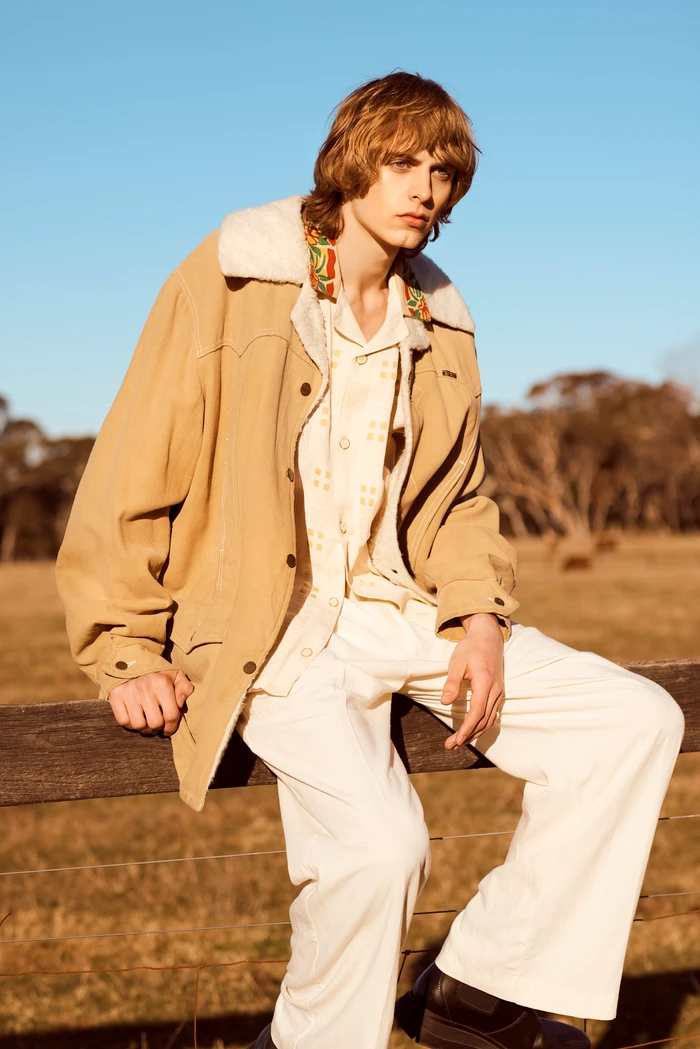 Young male model in tan shearling-collared jacket and white trousers sitting on a wooden fence outdoors.